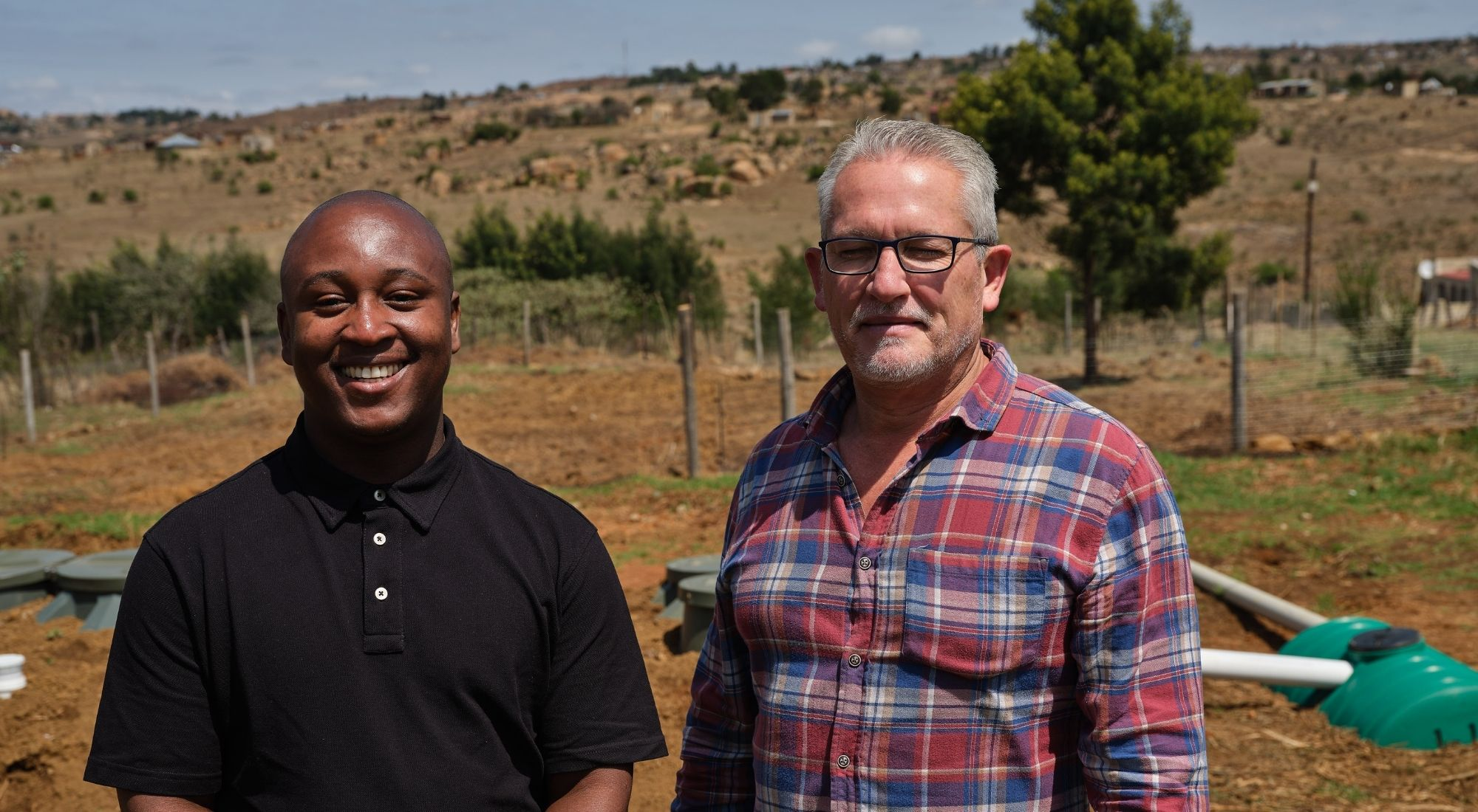 Two men standing beside biogas equipment in a rural landscape